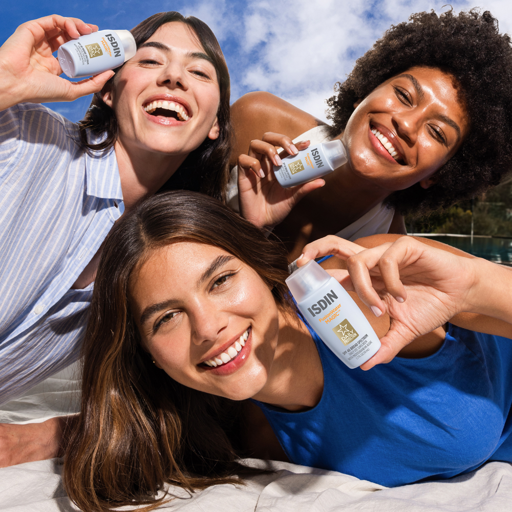 Three smiling women outdoors under a blue sky hold small Isdin sunscreen bottles, with water and a beach setting visible in the background.