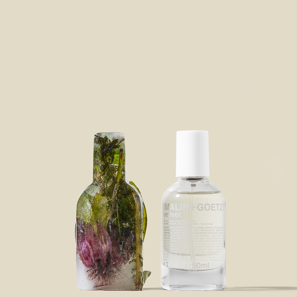 Malin & Goetz bottle with a white cap and printed label, and a sculptural clear bottle filled with green seaweed and purple sea life, with a small gold fish-shaped accent on a beige background.
