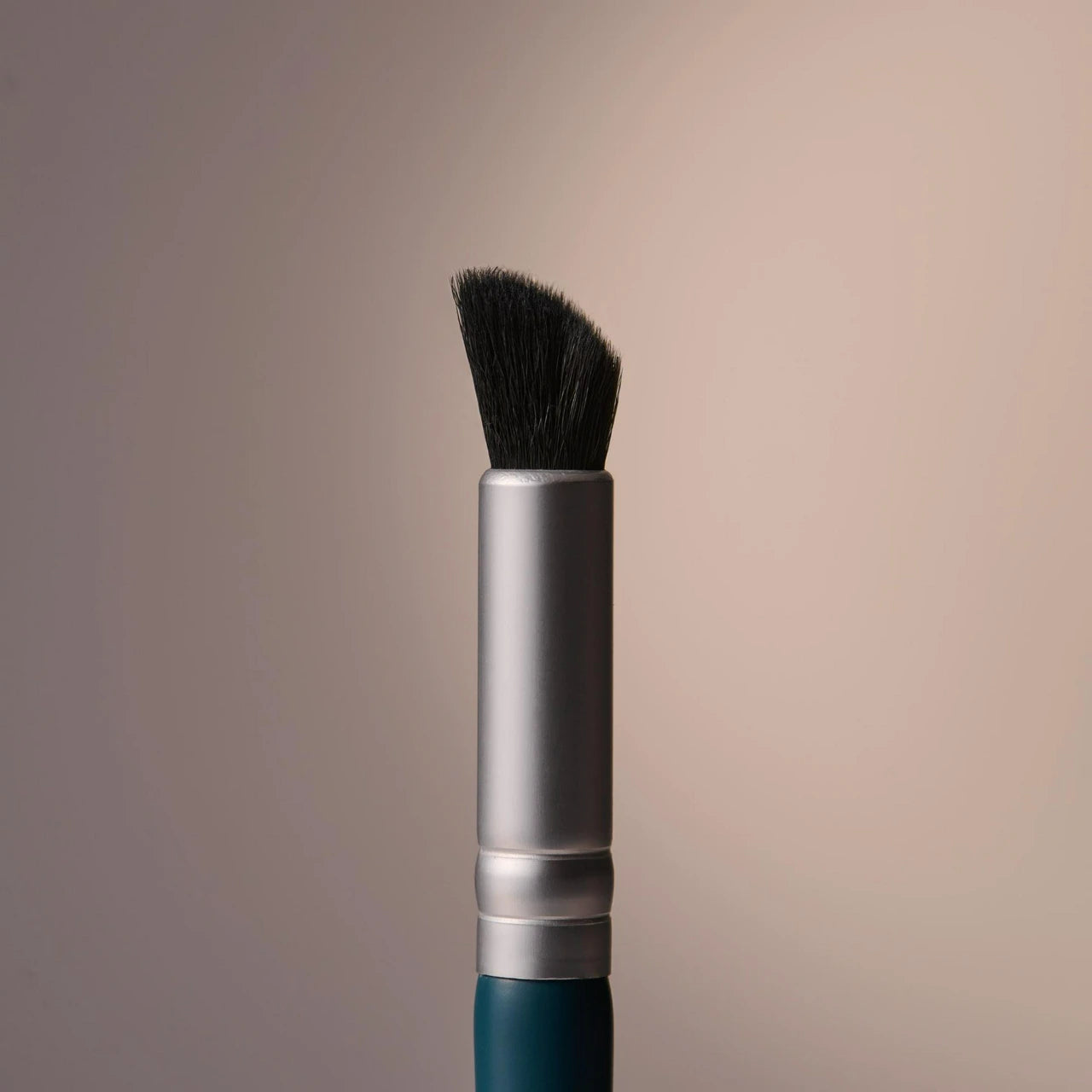 A close-up studio shot of a makeup brush with black bristles, a silver ferrule, and a dark teal handle, centered against a soft beige gradient background.