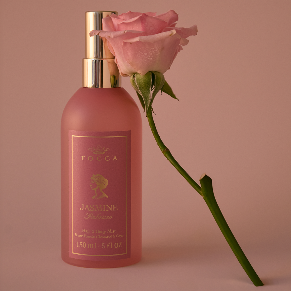 A bottle of TOCCA Jasmine Palazzo - Hair and Body Mist in a frosted pink bottle with a gold pump, styled with a single pink rose on a matching pink background.