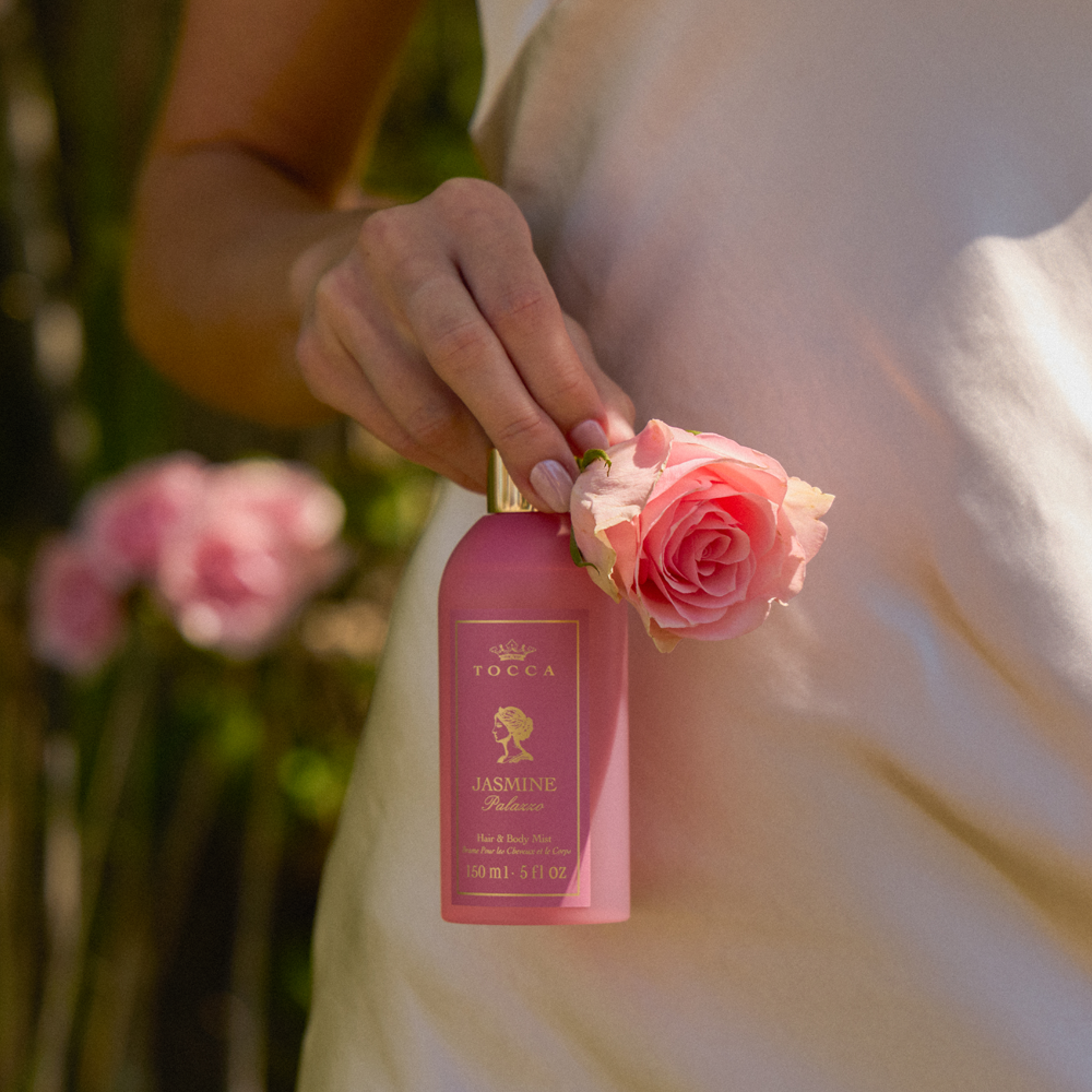 A person in a white dress holds a bottle of TOCCA Jasmine Palazzo - Hair and Body Mist next to a pink rose, with a soft-focus garden background.