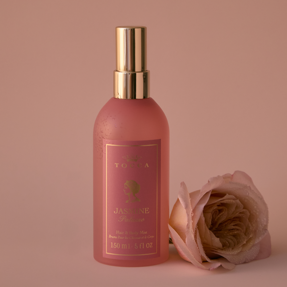 A bottle of TOCCA Jasmine Palazzo - Hair and Body Mist with a gold spray top, shown next to a matching pink rose on a solid pink background.