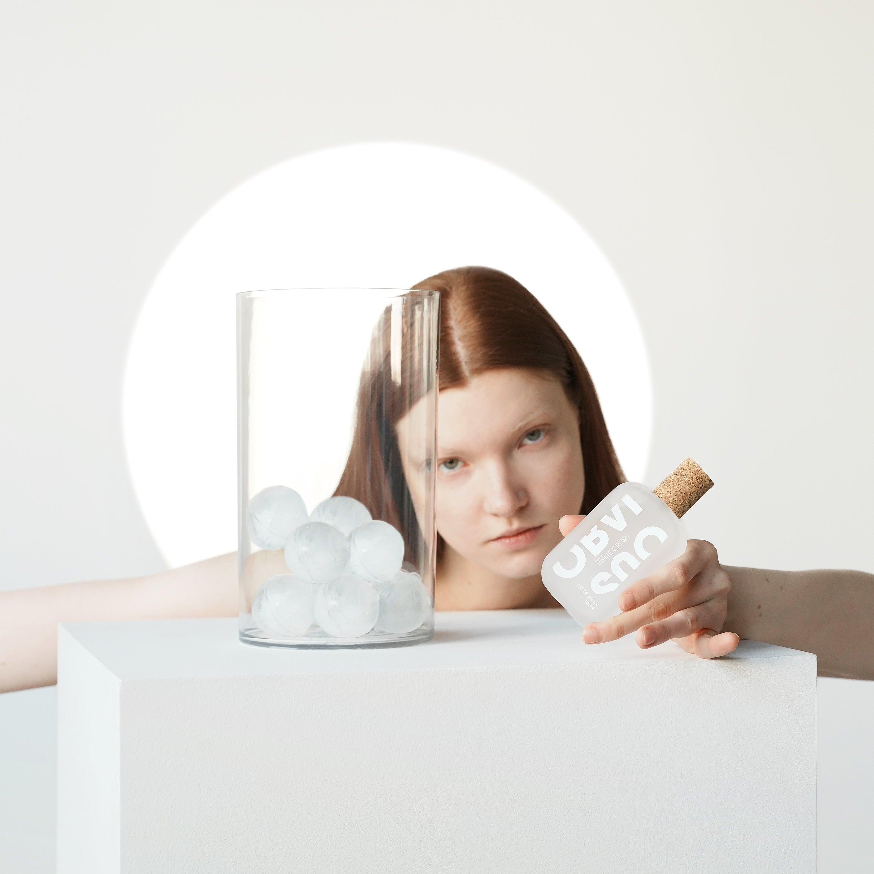 Person holding a perfume bottle next to a jar of ice cubes in front of a neutral background