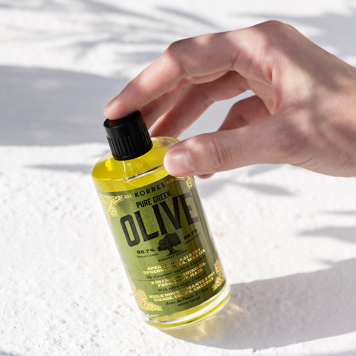 A hand holding a clear bottle of Korres Pure Extract Olive Oil against a bright white textured background with soft leaf shadows.