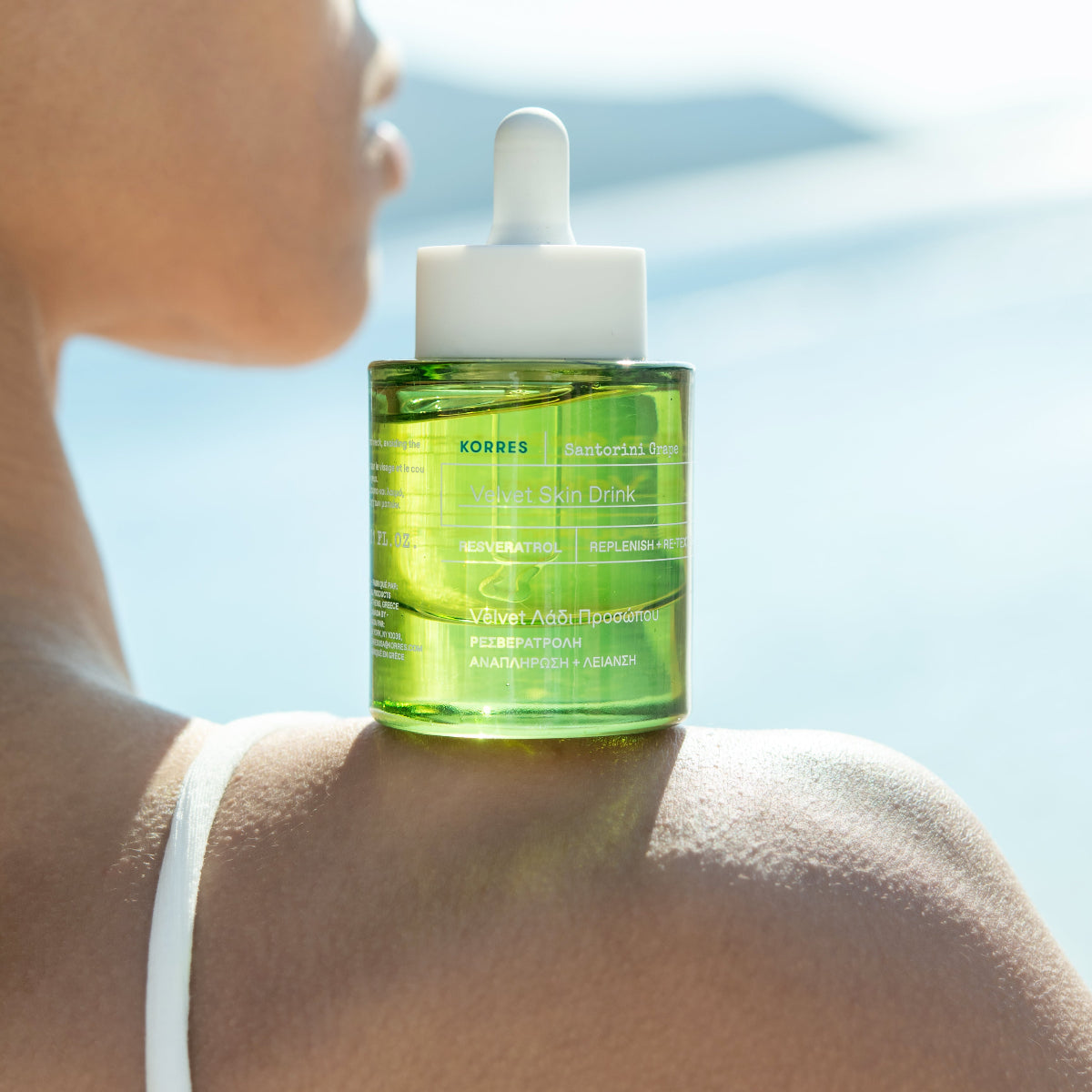 A bottle of Kores Velvet Skin Drink serum placed on a model's shoulder, with a blurred outdoor background.
