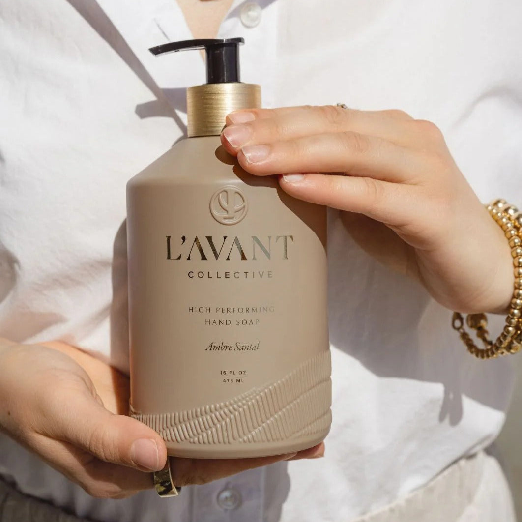 Person holding a beige bottle labeled 'L'AVANT Collective' against a white background