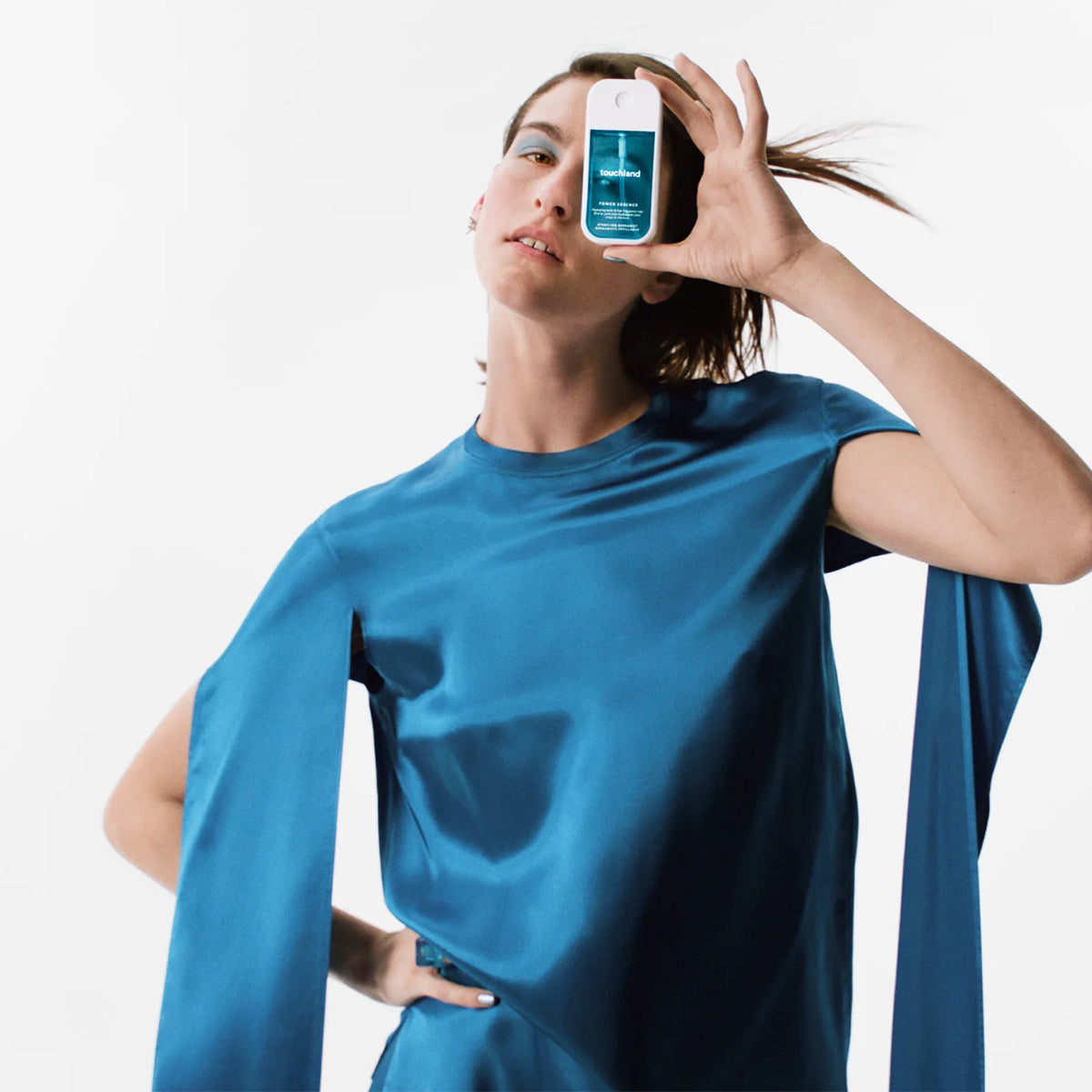 Person wearing a blue satin dress, holding Touchland Power Essence Hydrating Body & Hair Mist in front of their face, with a white background