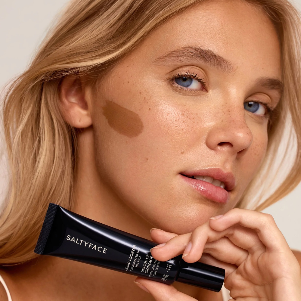 Woman applying bronzer with a Saltyface product on a neutral background