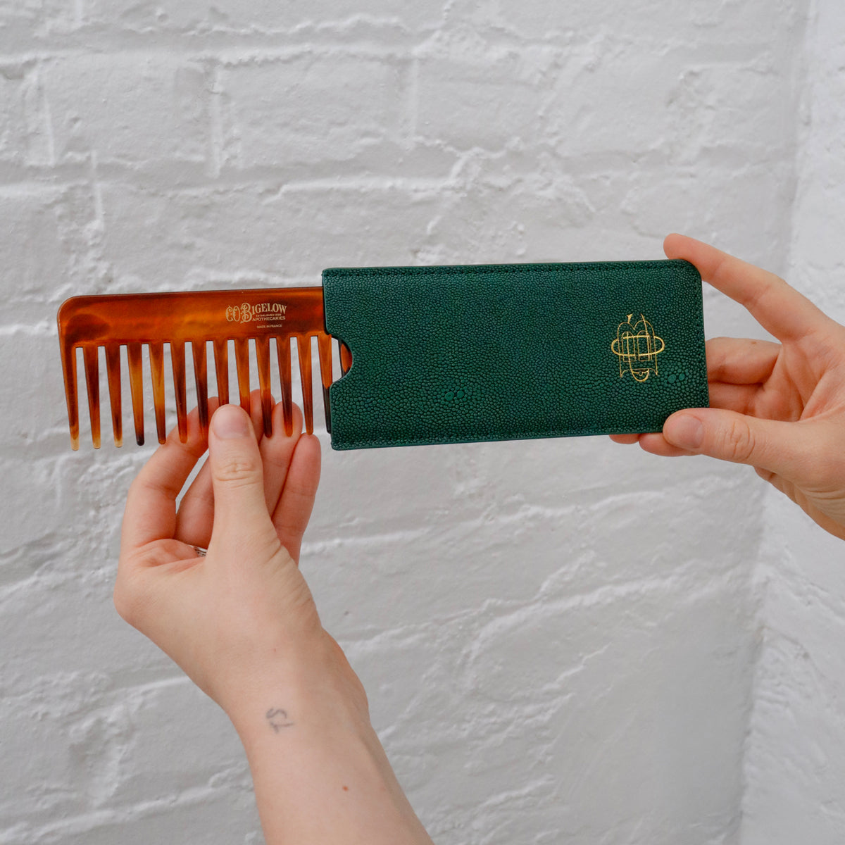 A person holds a wide-tooth comb with an attached dark green textured case against a white brick wall. The comb is amber-brown with white teeth, and the case has a gold logo.