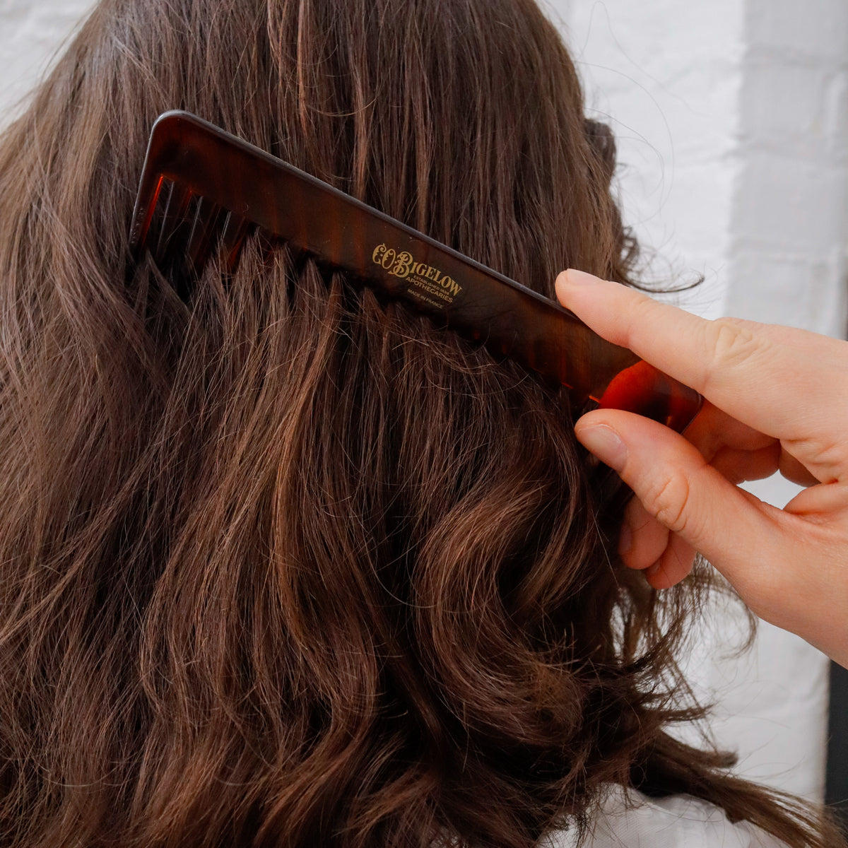 A close-up of a hand holding a dark brown tortoiseshell hair comb through wavy brown hair, with the brand name 'Bellew' printed in gold on the comb.