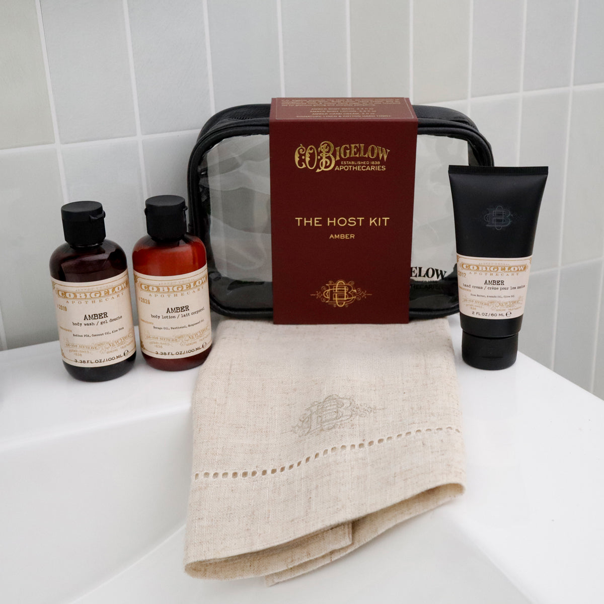 Bigelow Apothecary 'The Host Kit' amber set arranged on a white surface against a white tiled wall, including two amber bottles, a black toiletry tube, a dark red box, and a folded beige towel.