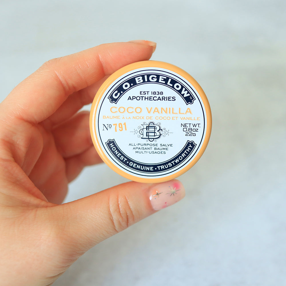 Hand holding a container of C.O. Bigelow Coco Vanilla all-purpose salve against a light background