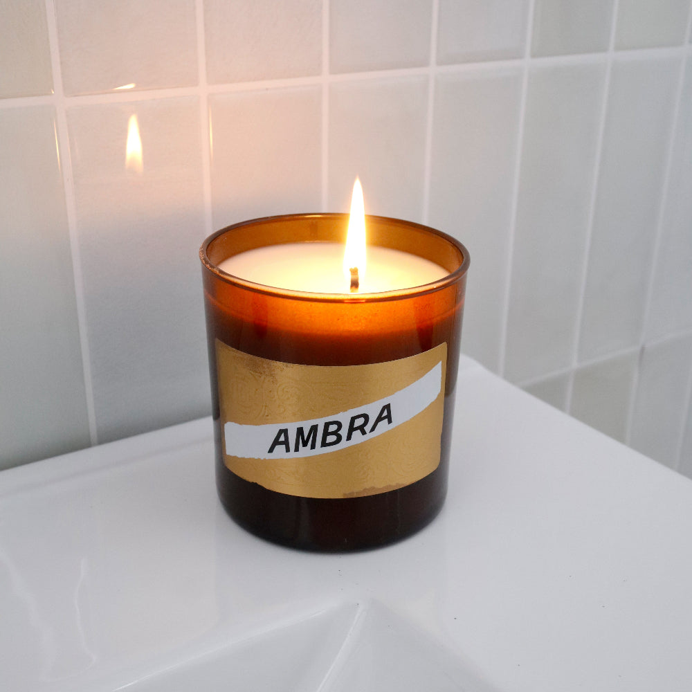 A lit amber glass candle jar with a gold label reading 'AMBRA' sits on a white bathroom counter against a white tiled wall.
