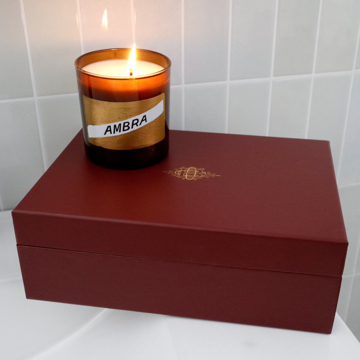 A lit amber glass candle labeled 'AMBRA' sits on top of a dark red rectangular box with a gold crest, placed against a light gray tiled wall.