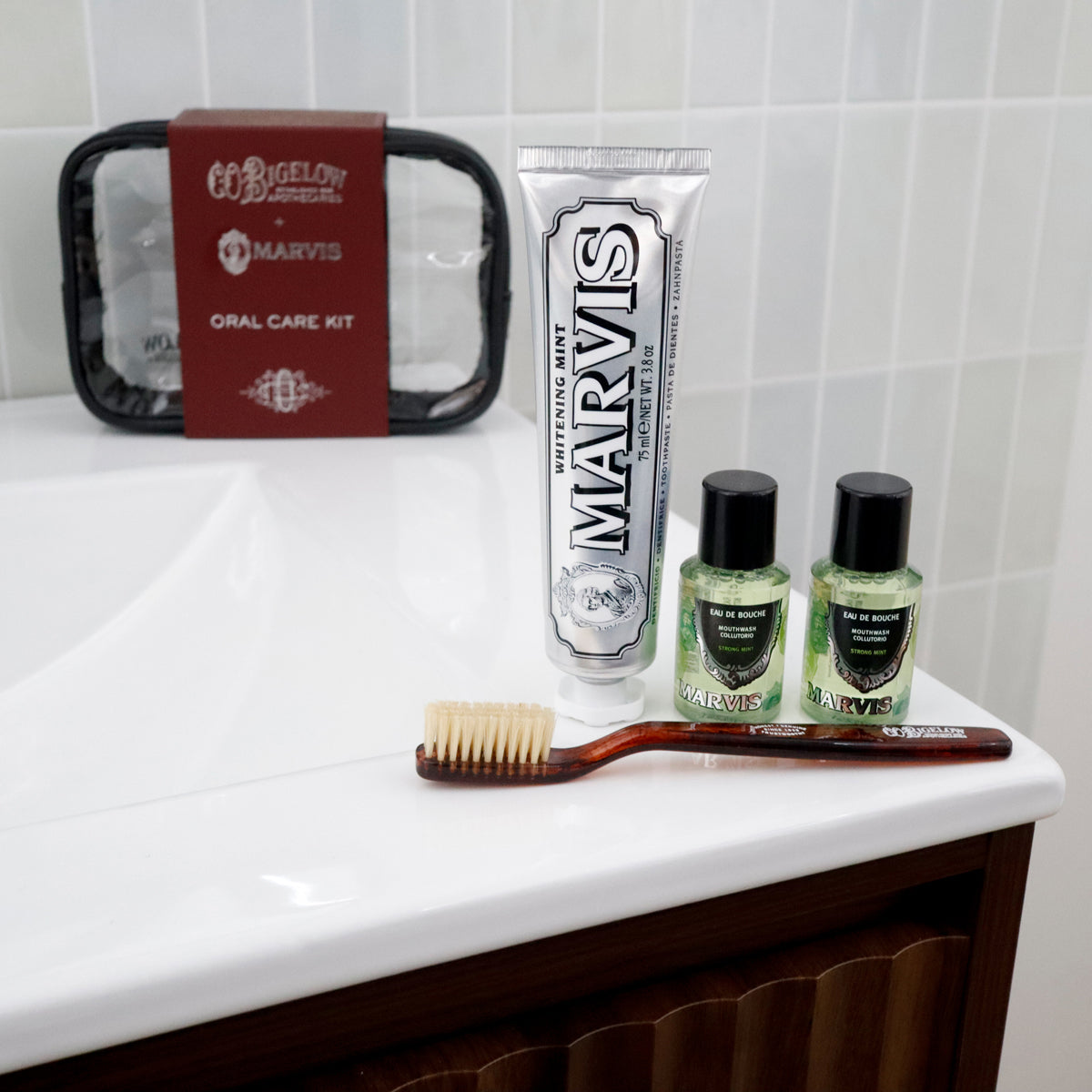 A Marvis oral care kit arranged on a white bathroom sink, including a tube of toothpaste, two small green bottles, a wooden toothbrush, and the black-and-brown kit pouch in the background.
