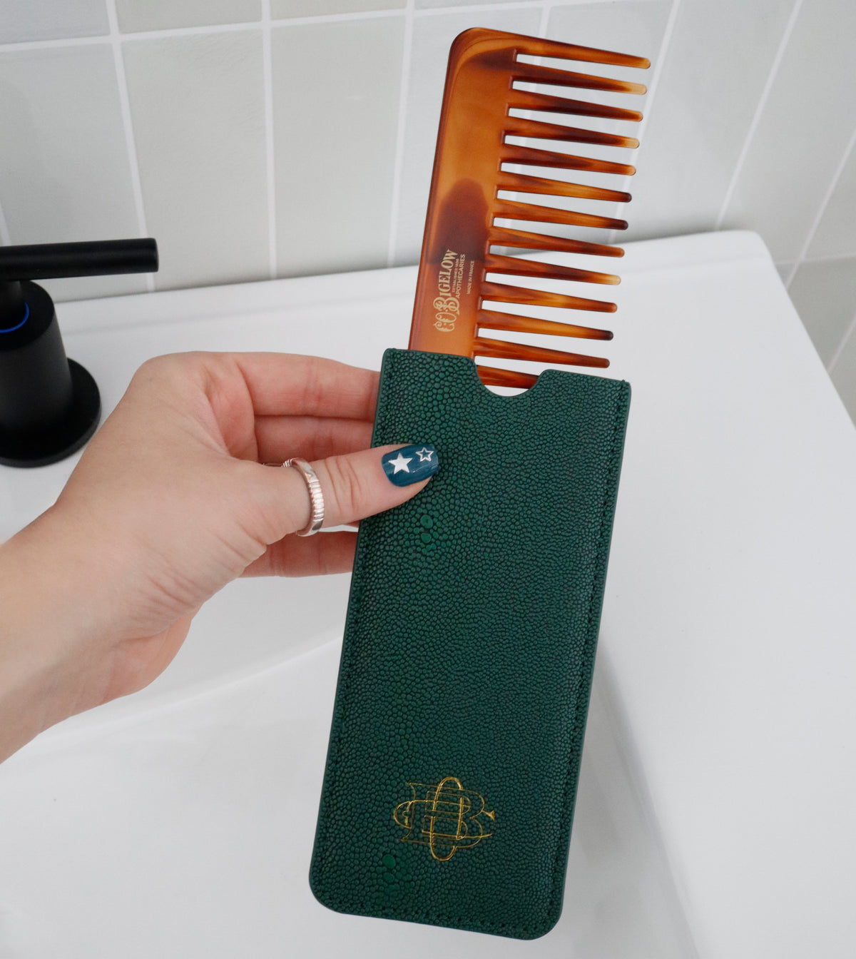 A hand holds a brown tortoiseshell hair comb partially inserted into a dark green textured travel case with a gold atom logo, set against a white tiled bathroom background.