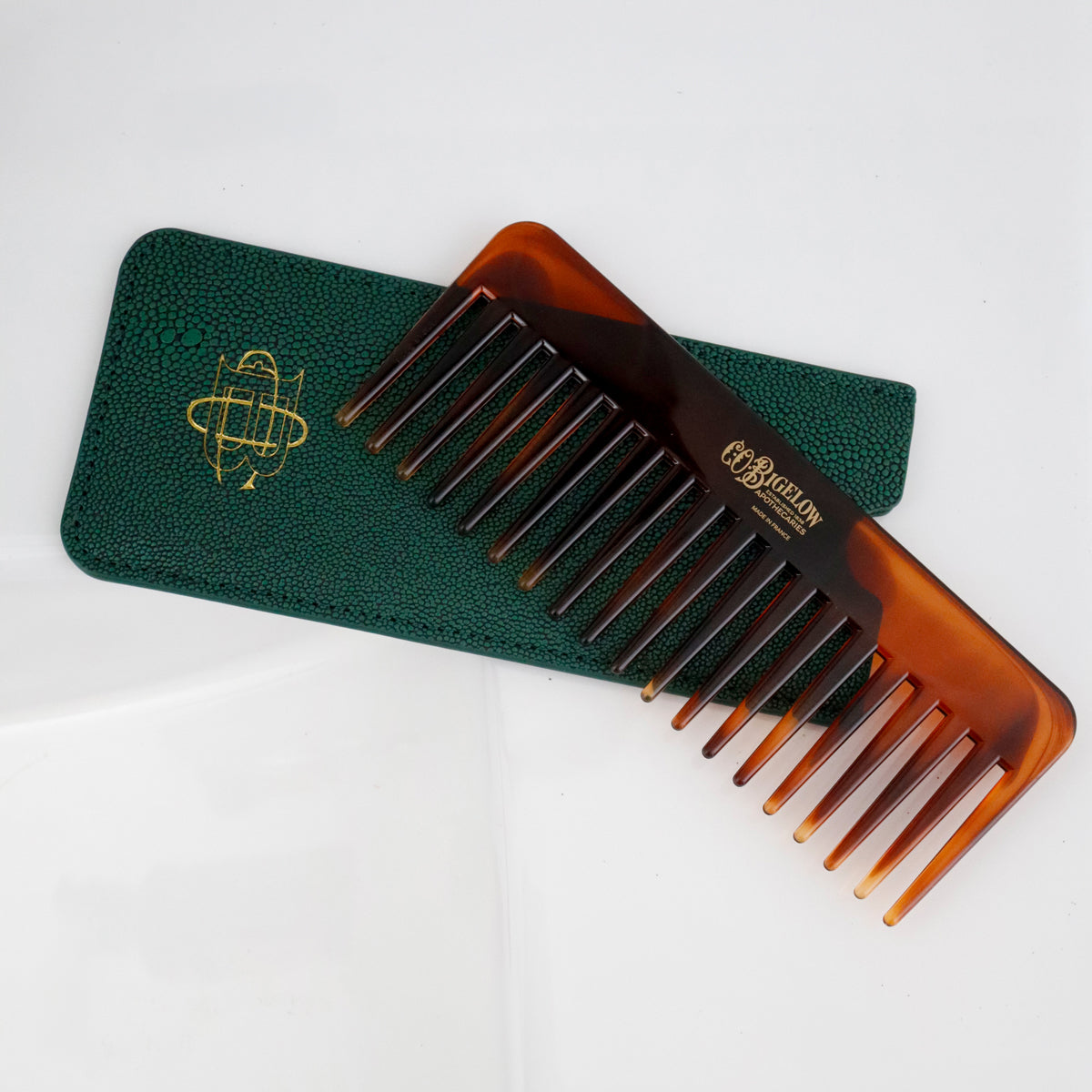 A brown tortoiseshell hair comb with a gold logo, resting diagonally on a dark green textured leather case with a gold emblem, on a clean white background.