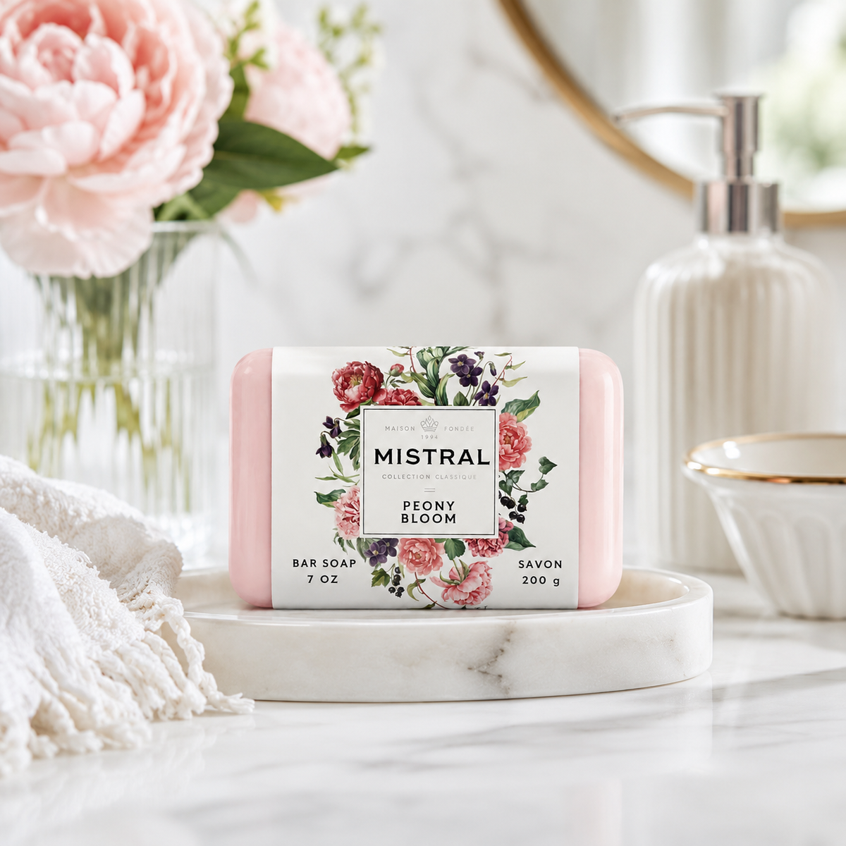 A pink bar of Mistral Peony Bloom soap sits on a white marble tray on a marble countertop. The scene is styled with a white towel, a glass vase of pink peonies, a ribbed white soap dispenser, and a small gold-rimmed bowl in a bright bathroom setting.