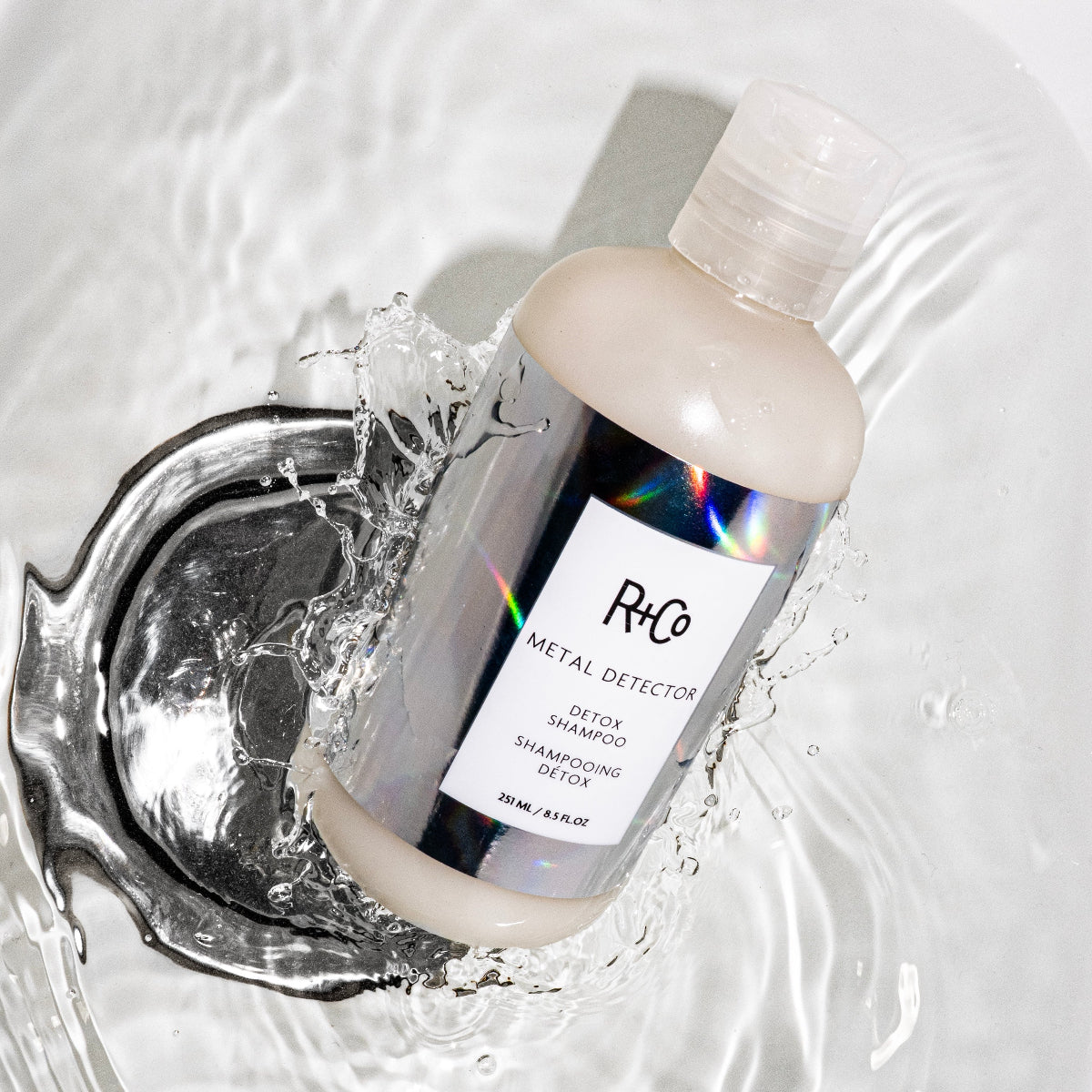 Bottle of R+Co metal detector shampoo and conditioner with water splash on a reflective surface