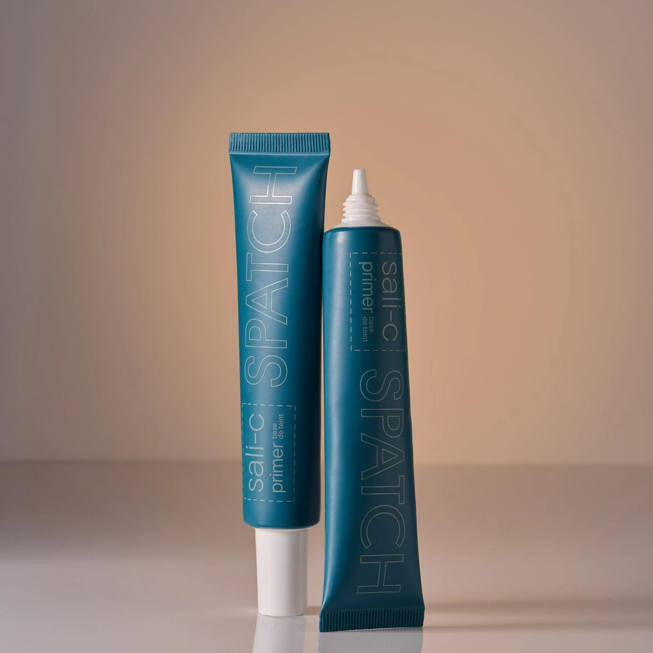Two teal-blue squeeze tubes of Sali-C Spatch primer stand upright on a light surface against a warm beige studio background. One tube is capped with a white flip-top, and the other has a pointed applicator tip.