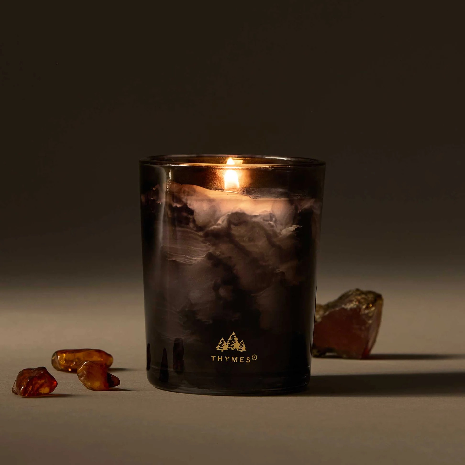 Lit candle in a dark glass container with 'Thymes' logo, surrounded by amber-colored stones on a dark background.