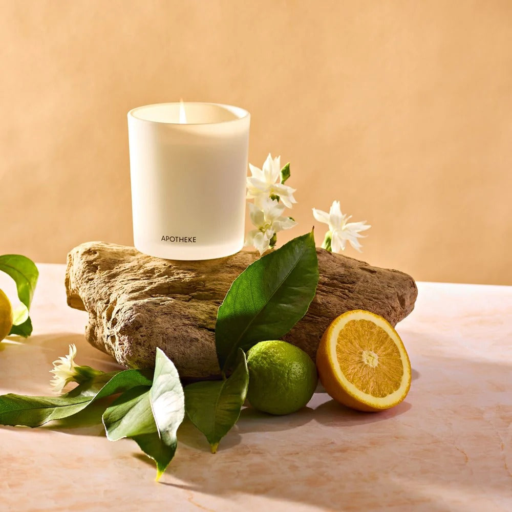 Orange Blossom Neroli Candle on a rock in front of a taupe background next to citrus fruits, white flowers and green leaves. 
