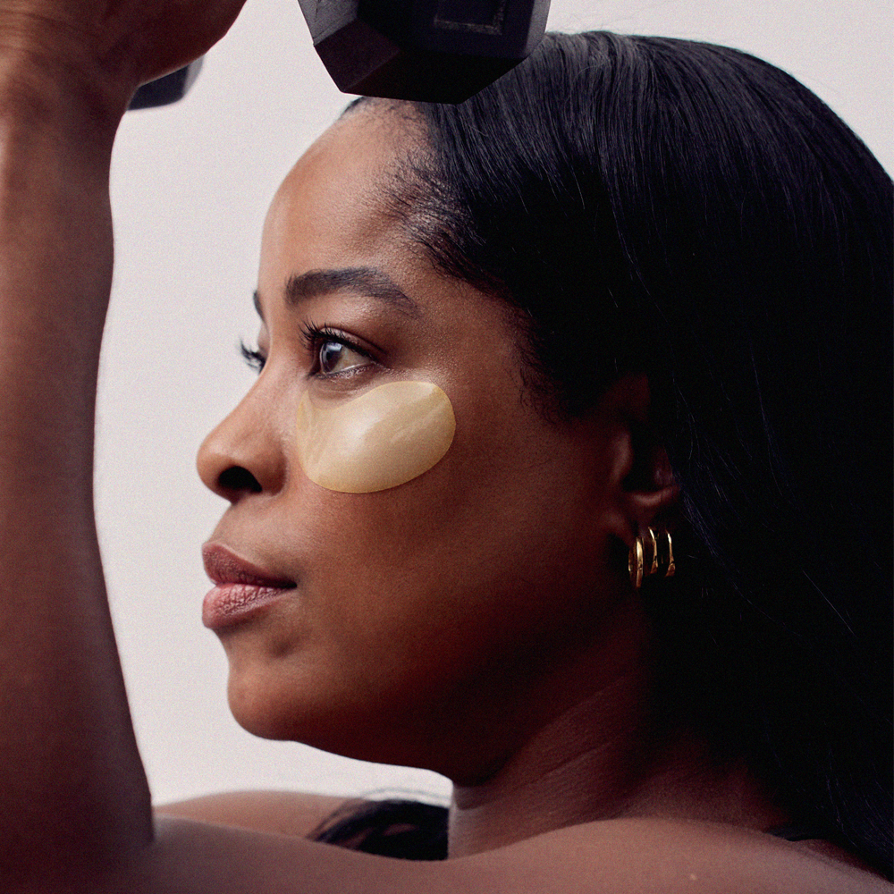 Close-up of a woman in profile with dark hair, wearing Radford Patch - the daily mask, gold hoop earrings, and holding a black dumbbell overhead against a plain light background.