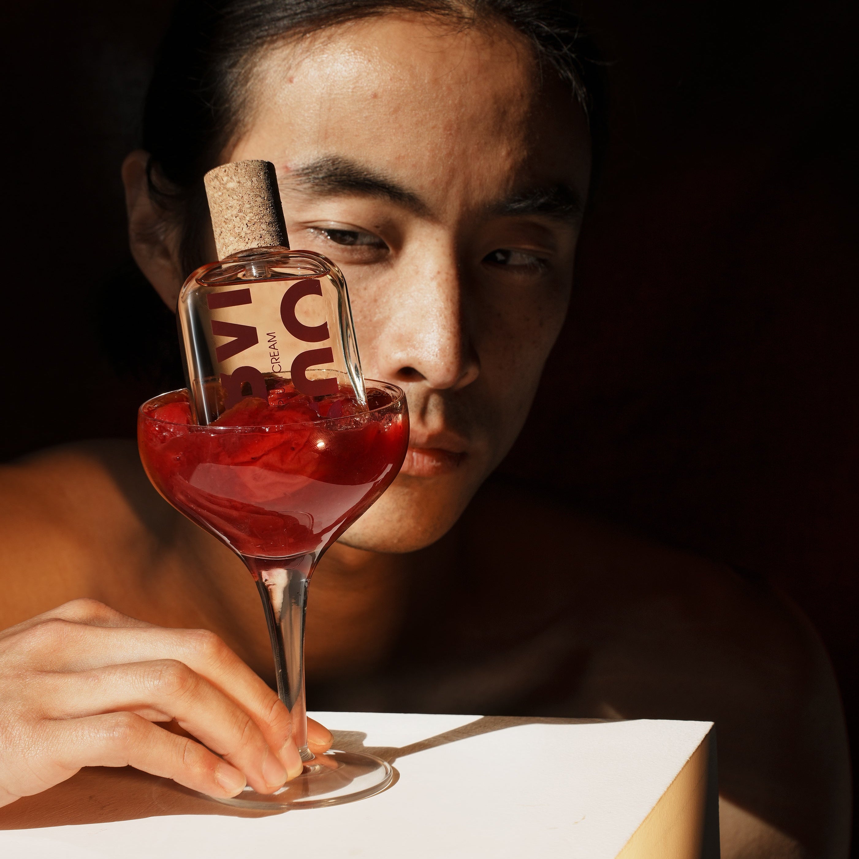Person holding a glass of red liquid  and a perfume bottle with a dark background