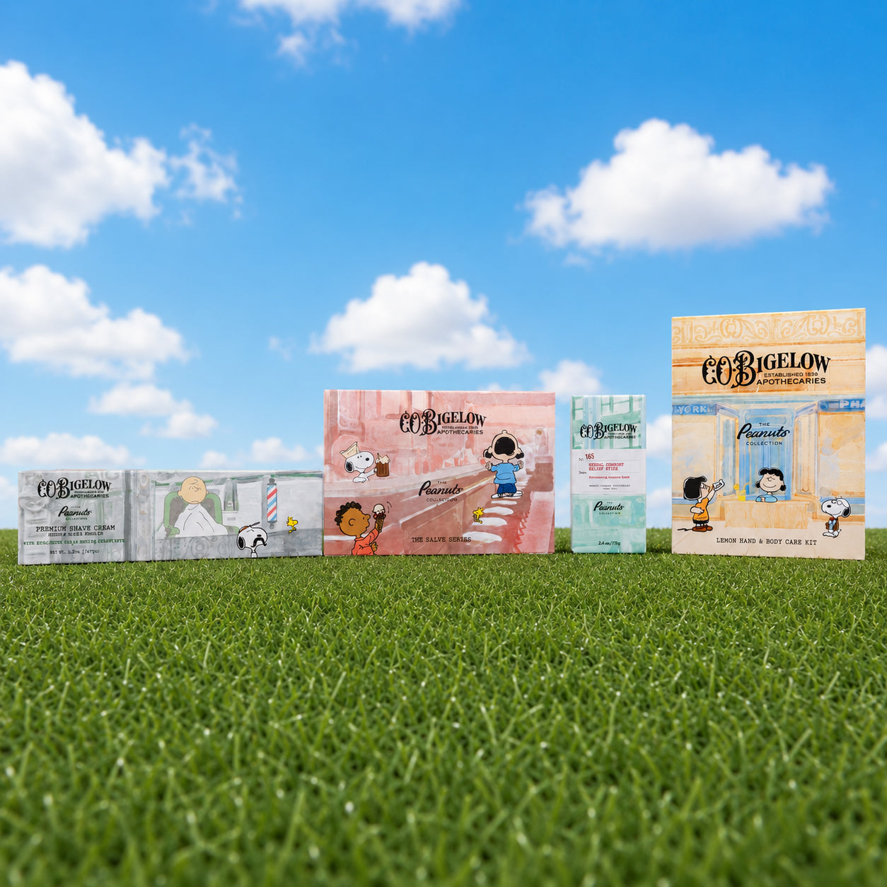 Five Co:Bigelow Apothecary product boxes arranged in a row on green grass under a blue sky with white clouds. The boxes feature cartoon illustrations from The Peanuts, including Snoopy, Woodstock, Charlie Brown, and other characters.