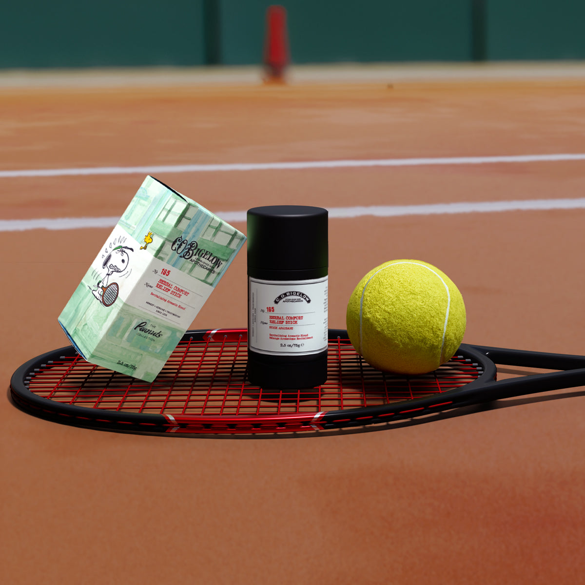 C.O. Bigelow Herbal Comfort Stick and its box displayed on a red tennis racket on an orange clay tennis court, next to a yellow tennis ball.