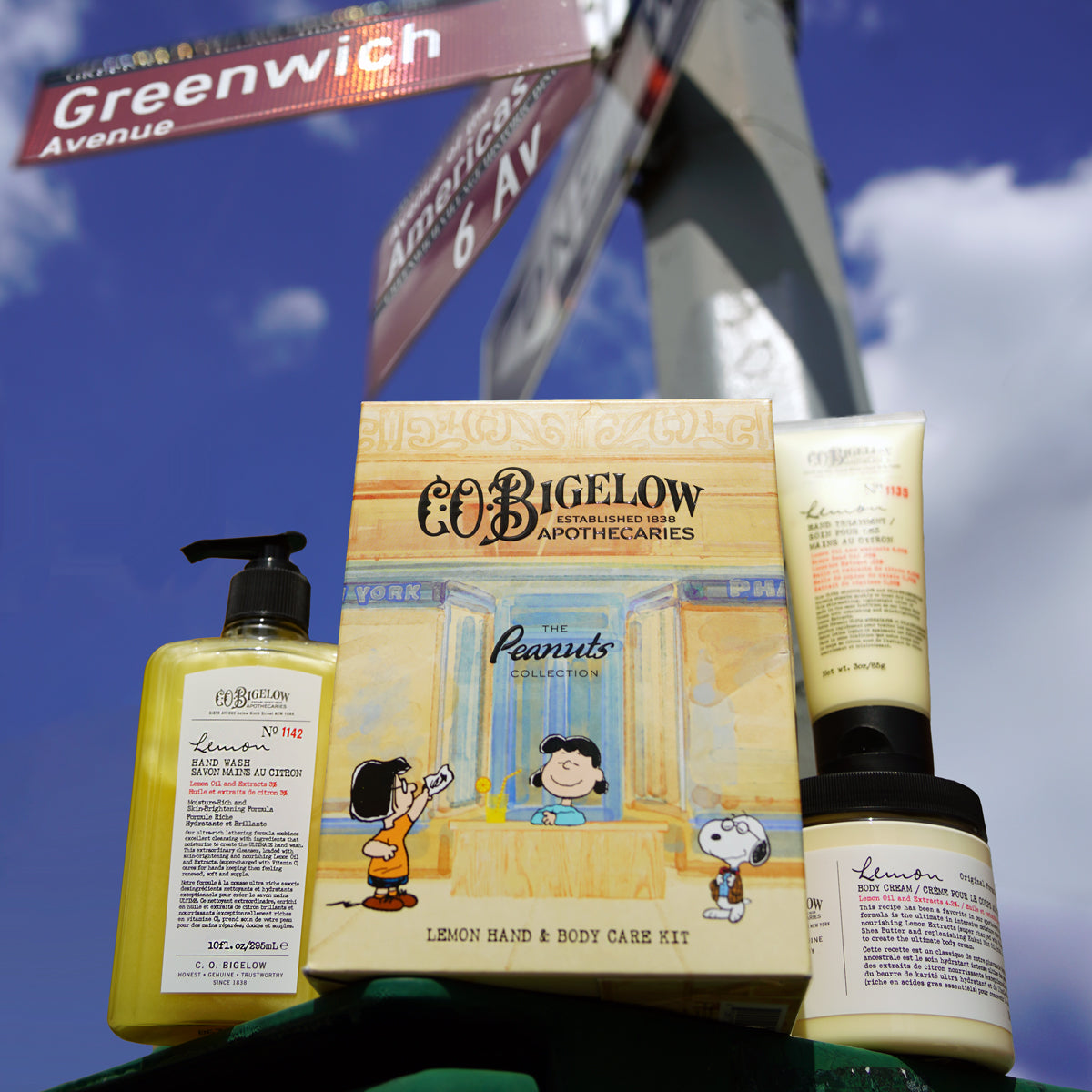Co: Bigelow Apothecaries The Peanuts Lemon Hand & Body Care Kit, featuring a yellow hand wash bottle, cream body lotion, and jar, displayed in front of a street sign and blue sky.