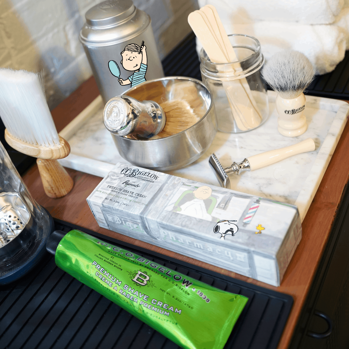 A styled grooming set on a wooden table, featuring a green tube of Bellow Men shave cream, a boxed shaving soap, wooden-handled brushes, a metal shaving bowl, a safety razor, and a silver tin canister.
