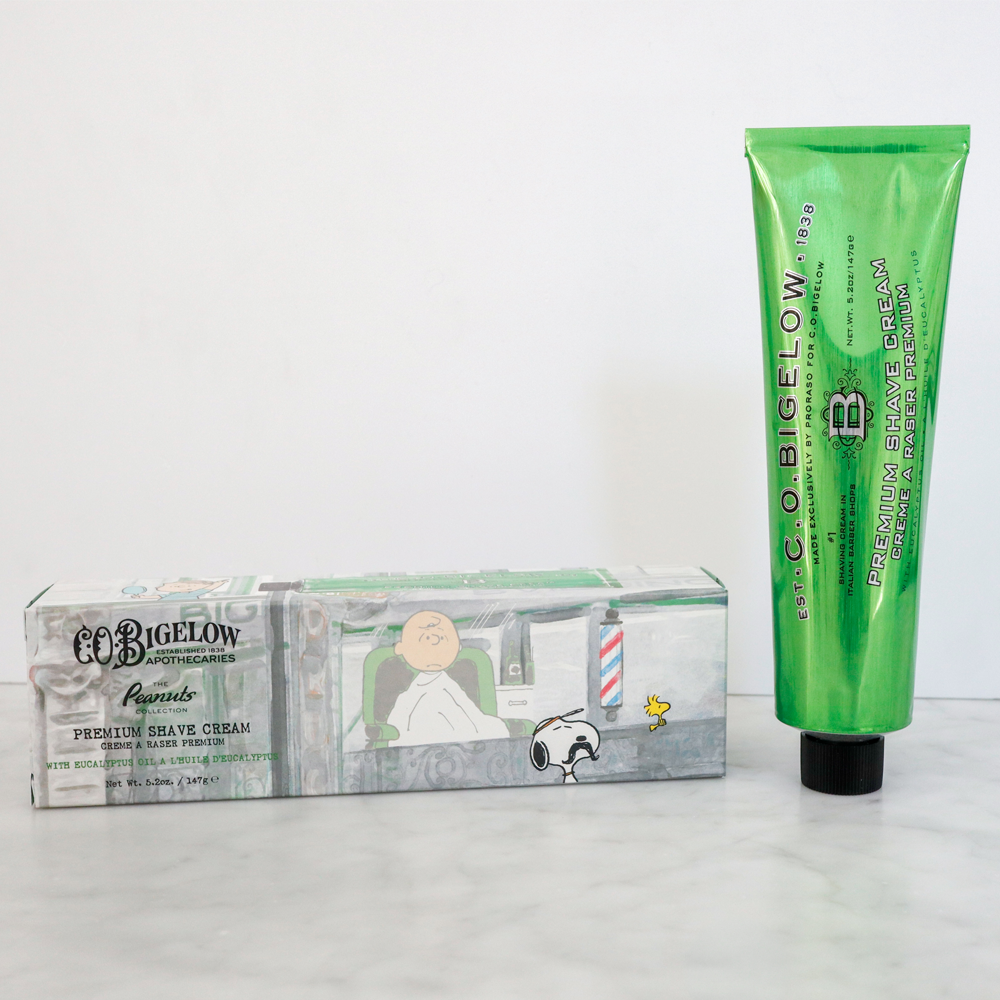 Peanuts® x C.O. Bigelow Premium Shave Cream and packaging on a marble surface in front of a white background