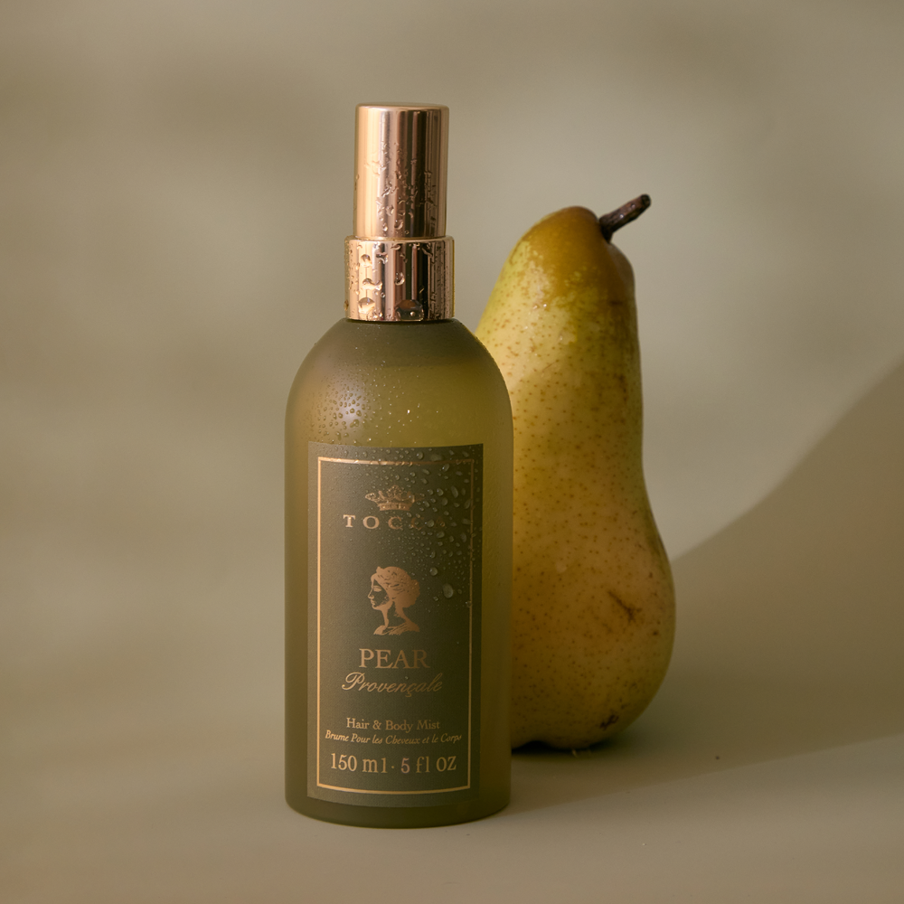 A bottle of TOCCA Pear Provencale - Hair and Body Mist stands next to a whole pear on a warm beige background.
