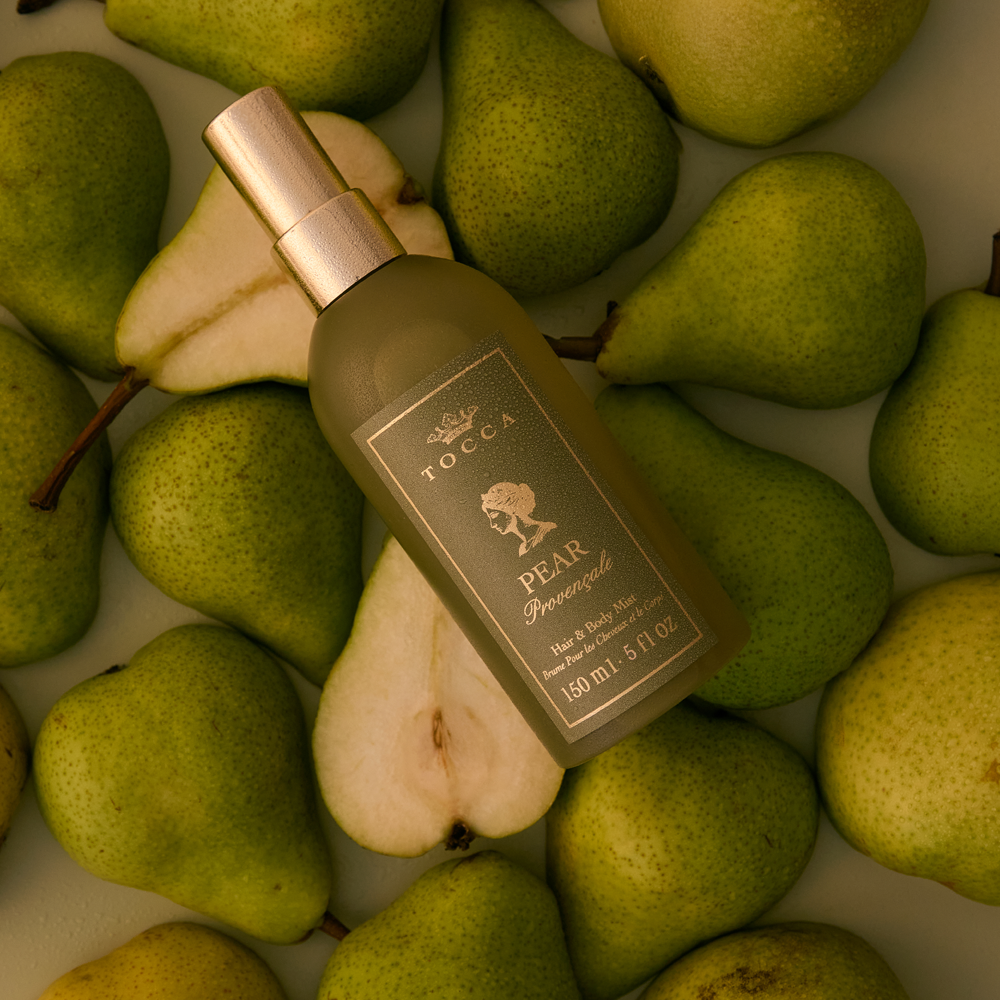 A bottle of TOCCA Pear Provencale - Hair and Body Mist lies diagonally on top of fresh green pears, including a sliced pear half showing the white flesh. The bottle is frosted olive green with a metallic gold spray nozzle and a matching gold label.