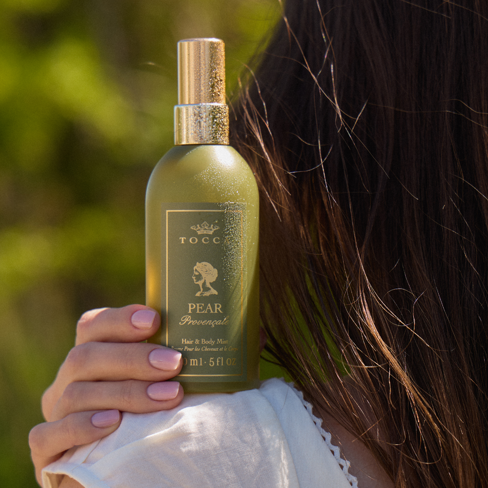 A woman holds a bottle of TOCCA Pear Provencale - Hair and Body Mist. The bottle is a muted olive green with a shiny gold spray nozzle and a gold label.