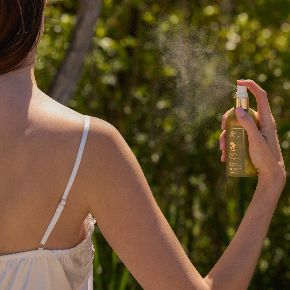 A woman outdoors holds a bottle of TOCCA Pear Provencale - Hair and Body Mist, spraying mist toward her face, with a blurred green natural background.