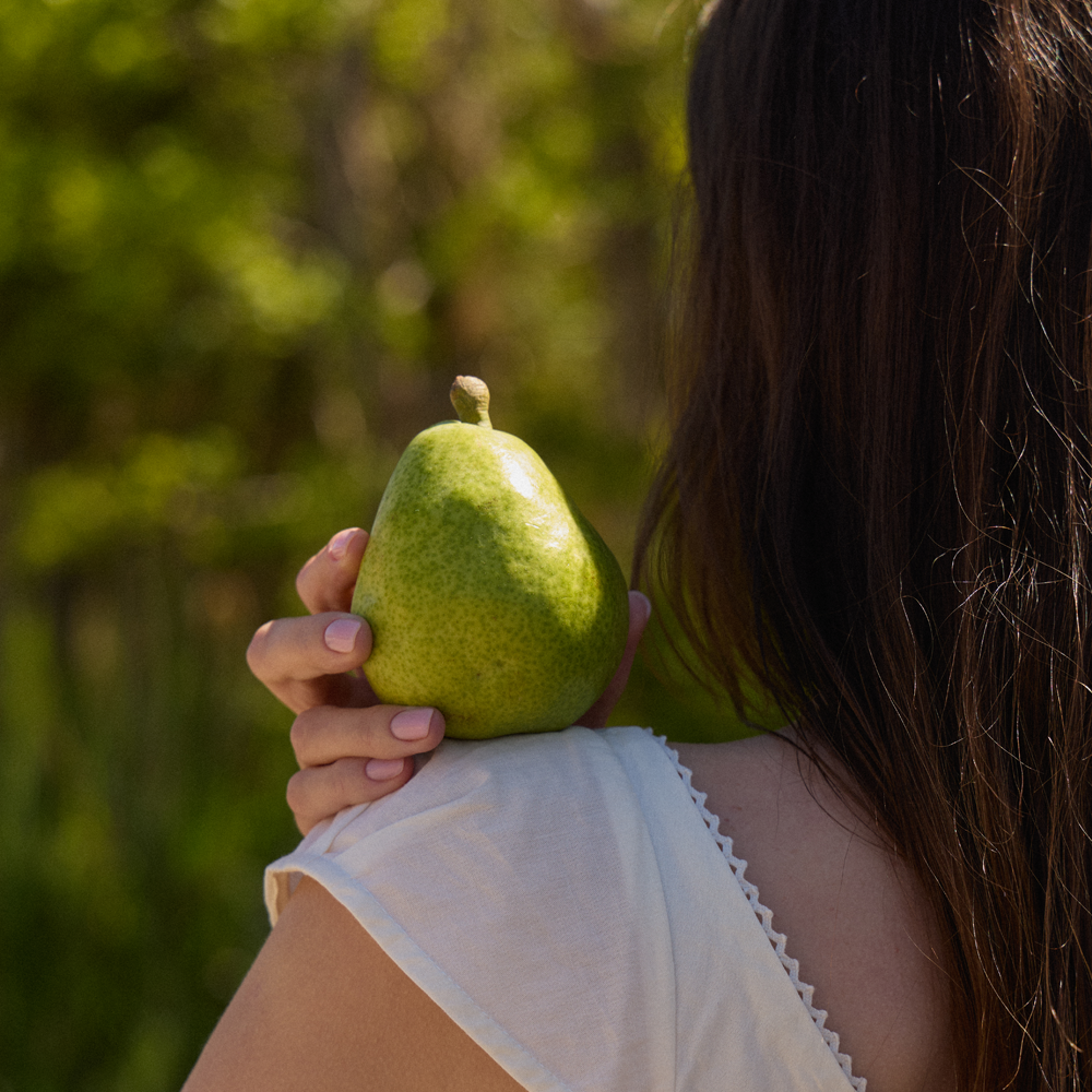A woman in a white top holds a green pear against her shoulder, photographed outdoors with a blurred green forest background.