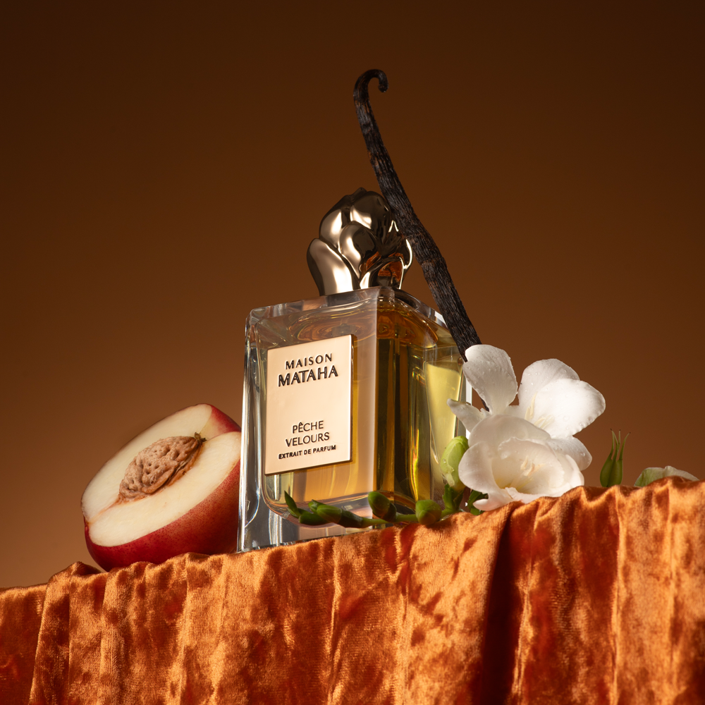 Maison Mataha Pèche Velours perfume bottle styled on an orange velvet surface with a sliced peach, white flowers, and a vanilla pod against a warm brown background.