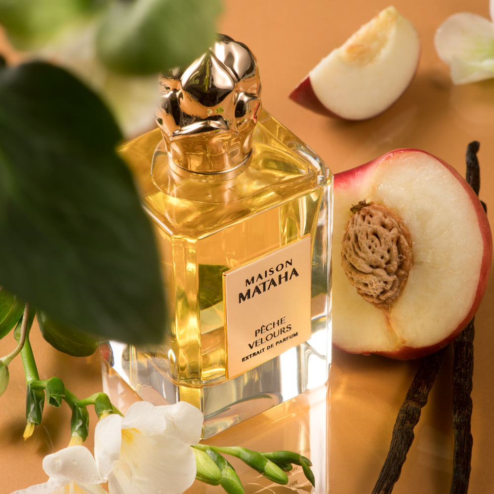 Maison Mataha Pèche Velours perfume bottle with a gold cap, styled with sliced peaches, vanilla beans, and white flowers on a warm tan background.