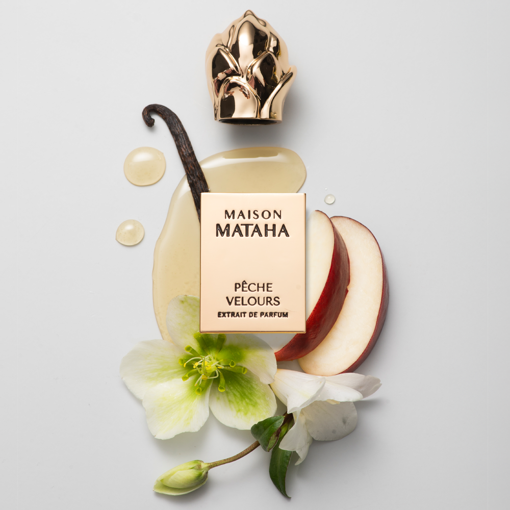 Maison Mataha Pèche Velours extrait de parfum perfume bottle with a gold cap, styled with apple slices, a vanilla bean, and white flowers on a light gray background.