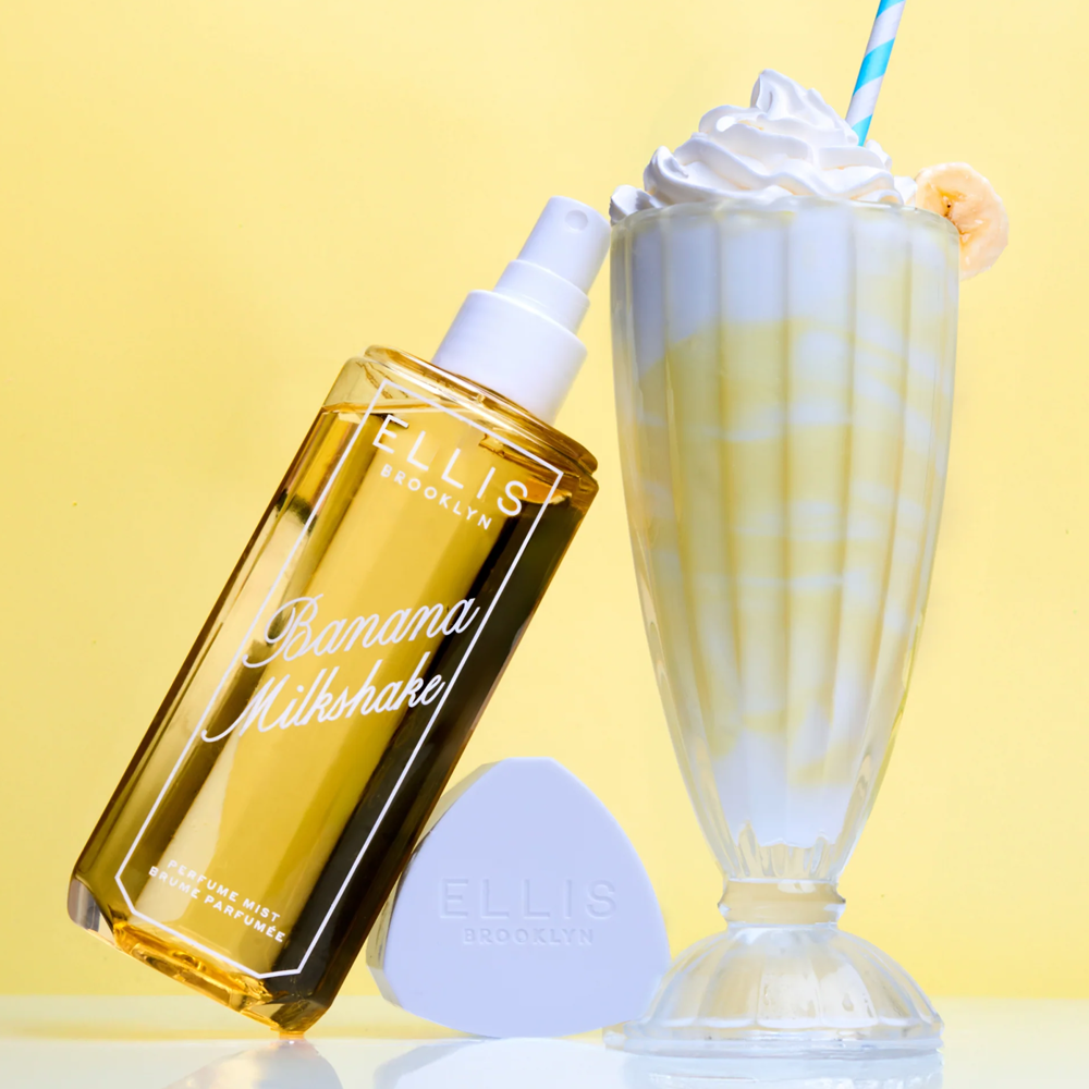 Yellow bottle labeled 'Banana Milkshake' by Ellis Brooklyn next to a glass of banana milkshake with whipped cream on a yellow background.