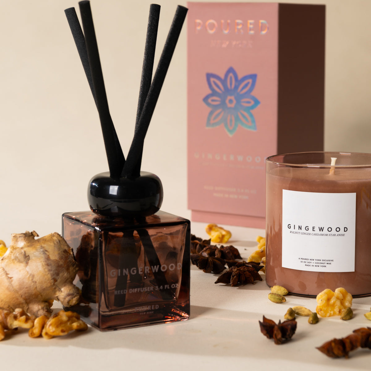 A gingerwood-scented reed diffuser bottle with black reeds in the foreground, a matching candle jar with a white label, and a pink branded box in the background, styled with ginger root, walnuts, and spices on a warm beige surface.