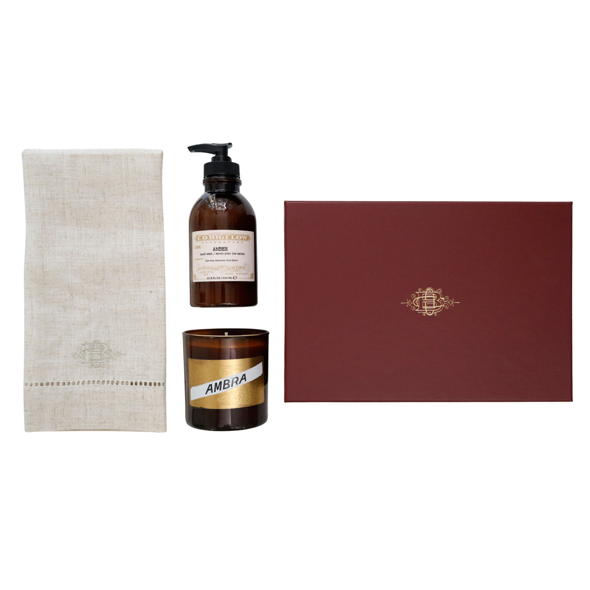 A gift set featuring a beige linen towel, an amber glass bottle with a black pump dispenser, a matching amber glass candle jar with a gold label, and a dark red rectangular box with a gold crest, all arranged on a white background.