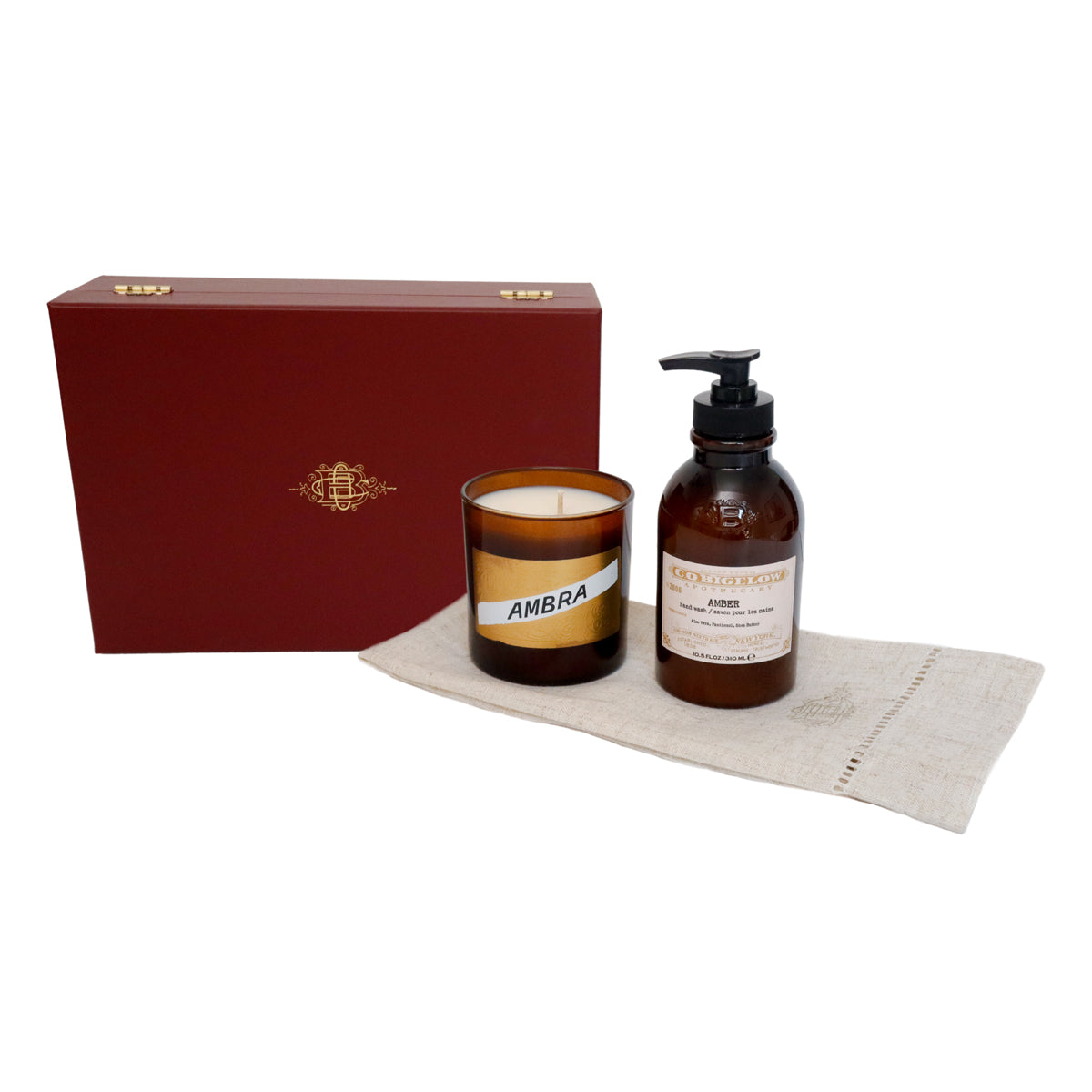 A gift set featuring an amber glass candle jar labeled 'AMBRA' and an amber pump bottle with a black dispenser, placed on a beige cloth in front of a dark red presentation box with gold detailing.