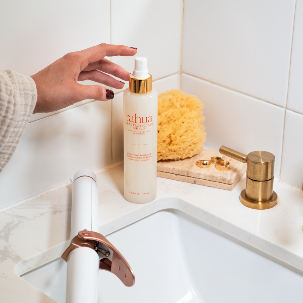 Hand holding a bottle of Rahua hair product next to a sink with a sponge and soap dispenser.