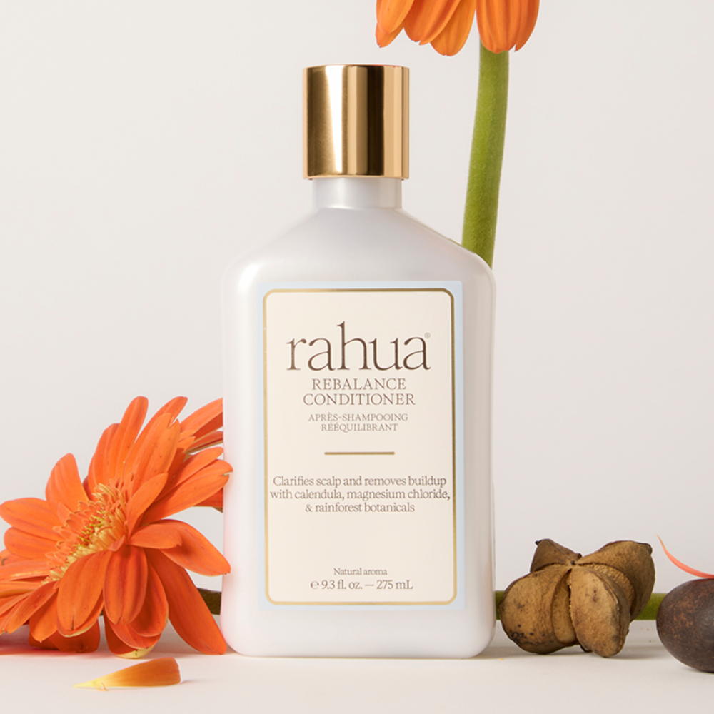 A bottle of rahua REBALANCE CONDITIONER stands centered on a light neutral background, styled with orange flowers and botanical elements around it. The bottle is frosted white with a metallic gold cap and a cream label with black text.
