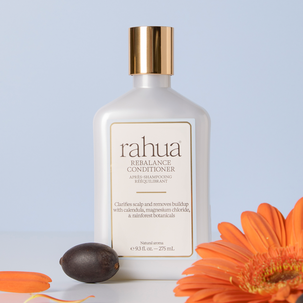 A bottle of Rahua Rebalance Conditioner after shampooing, styled with a dark brown seed and orange flower petals on a light blue background.