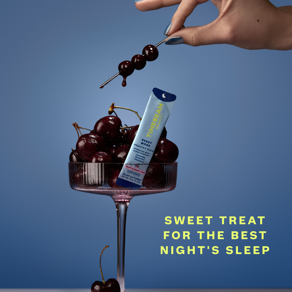A hand holding a toothpick with cherries over a glass with cherries and one packet of Timebeam Beauty - PM Reset Mode Healthy Pixie in front of a light blue background