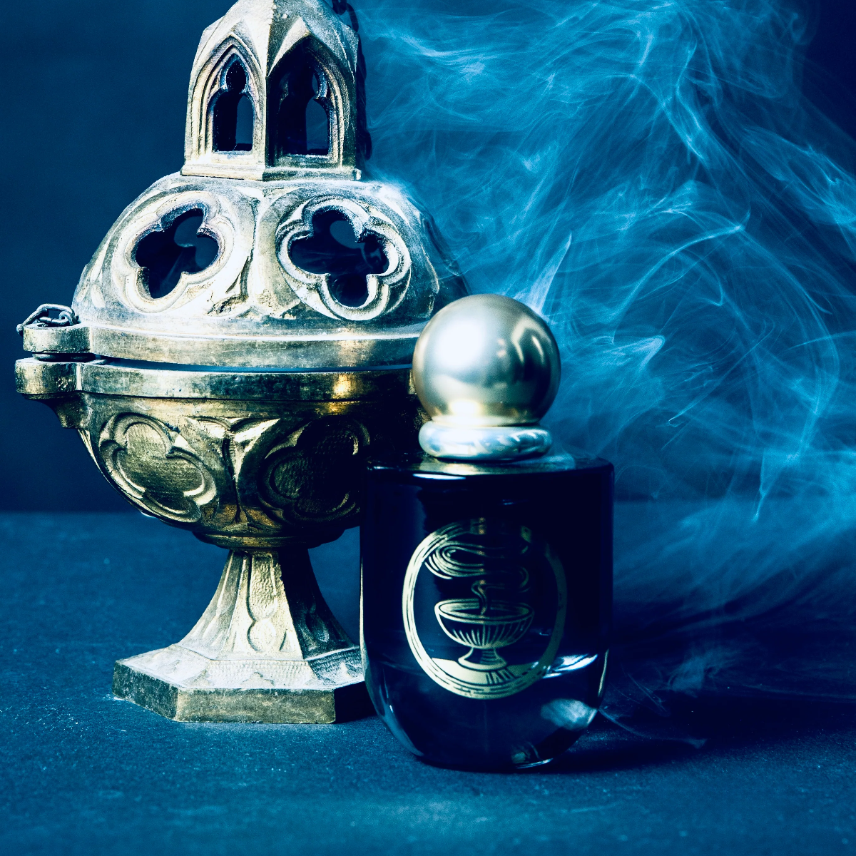 A dark glass perfume bottle with a metallic silver spherical cap and ornate emblem, positioned next to an antique silver incense burner with blue smoke drifting behind it on a dark blue background.